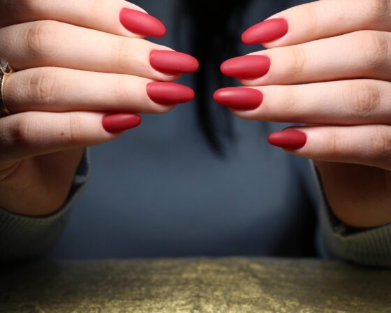 Two hands with matte red nails posed above a gold textured surface, with rings on two fingers