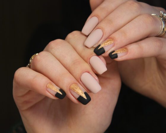 Close view of overlapping hands on a dark background, showing medium matte nude nails with black tips and gold glitter panels on accent nails