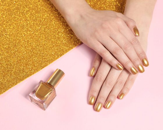 Two hands resting beside a gold glitter sheet and a bottle on pale pink, showing short metallic gold nails