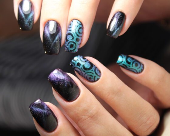 Tight close-up of two hands against a dark blurred background, showing dark navy-violet shimmer nails with turquoise circle patterns on accent nails