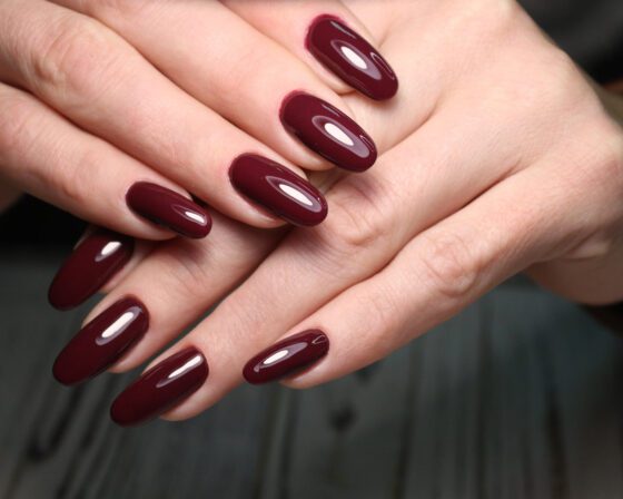 Tight view of crossed hands against a dark blurred background, showing medium-length nails in a single glossy deep burgundy shade