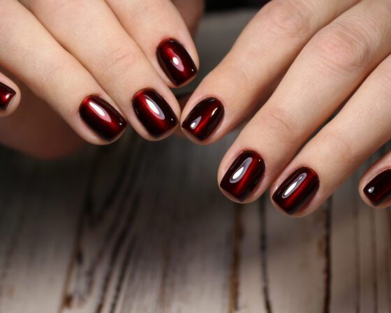 Close crop of two hands posed around a dark central background, showing short squared nails in black cherry with a luminous red centre