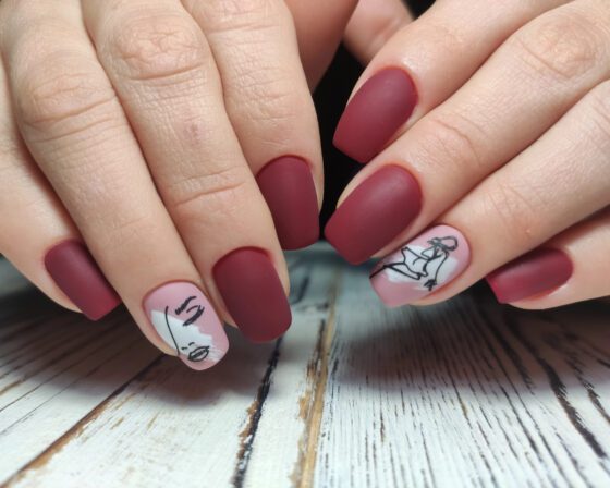 Multiple hands close together above pale wood, showing matte berry-red nails with pale pink accents carrying black line drawings
