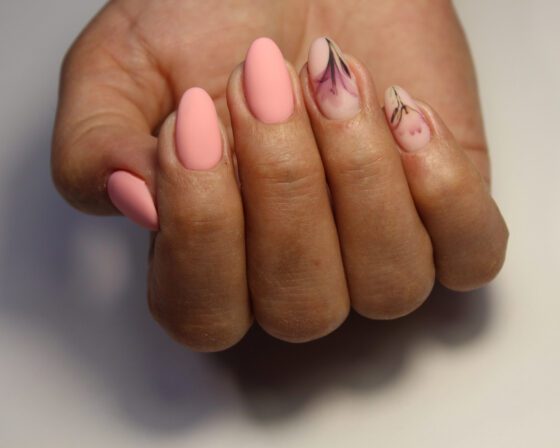 Close-up of one hand against a warm gray background, showing matte peach almond nails with fine dark botanical sketches on sheer accent nails