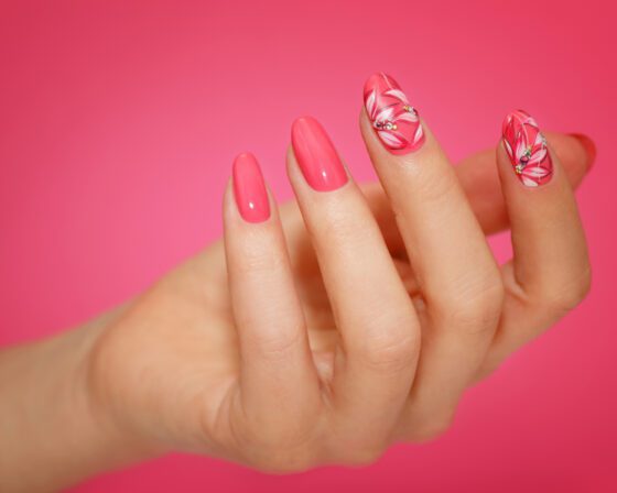 Close-up of one hand against a saturated pink studio background, showing glossy coral pink almond nails with floral line art on accent nails