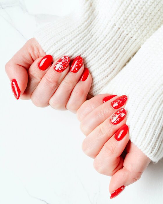 Two hands in white knit sleeves against a bright pale background, showing bright red nails with white snowflakes and fine sparkle on accent nails