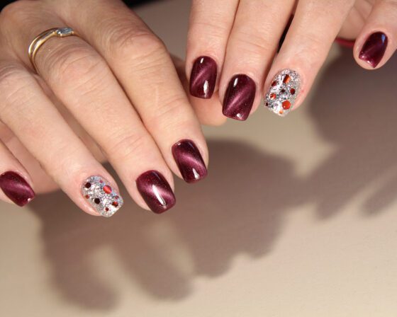 Close-up of multiple hands against a bright neutral background, showing wine red nails with a luminous sheen and a silver glitter accent set with small red and gold stones
