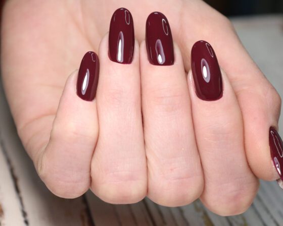 Close-up of one hand on pale wood with a dark blurred background, showing medium-length nails in a single glossy deep burgundy shade