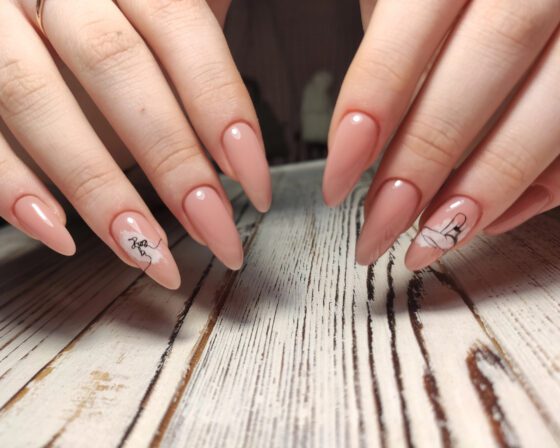 Close-up of overlapping hands above pale wood, showing long glossy peach-nude almond nails with small black line drawings on accent nails