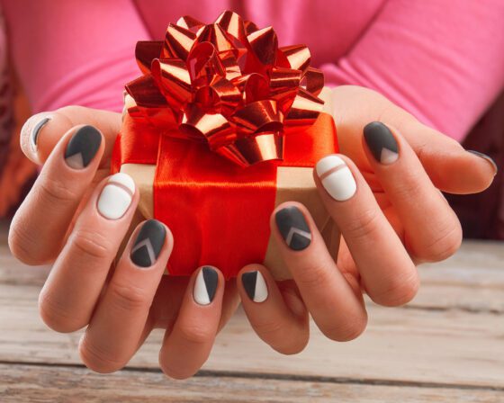 Two hands holding a red gift box with a ribbon bow over pale wood, showing short nails with charcoal, white and grey geometric colour blocks