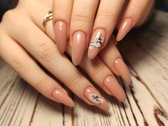 Macro view of one hand against pale wood, showing long glossy peach-nude almond nails with small black line drawings on accent nails