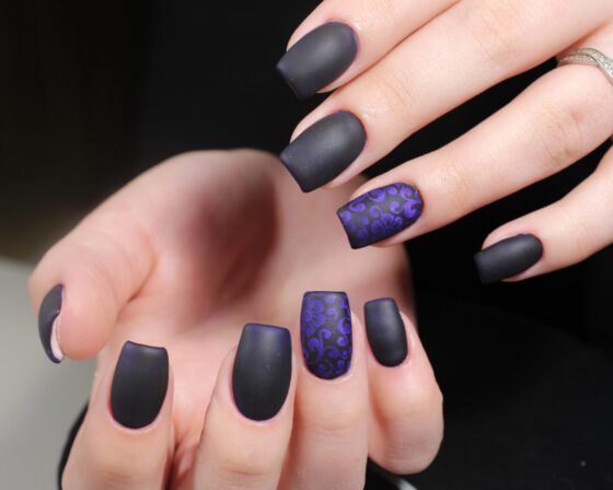 Close-up of two hands against a black background and pale tabletop, showing matte charcoal nails with a patterned purple accent nail
