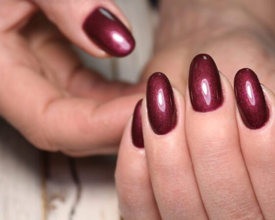 Extreme close-up against a softly blurred gray-toned surface, showing medium oval nails in a deep wine red with a fine metallic shimmer