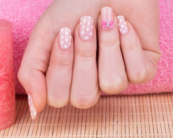 Centered close-up of one raised hand against pink and woven textures, showing pale blush nails with white polka dots and small pink jewelled accents