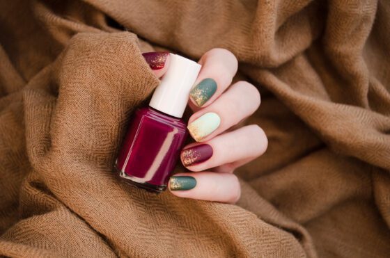 Close-up of hand against brown knitted fabric, showing short matte nails in forest green, wine and cream with gold glitter at the tips
