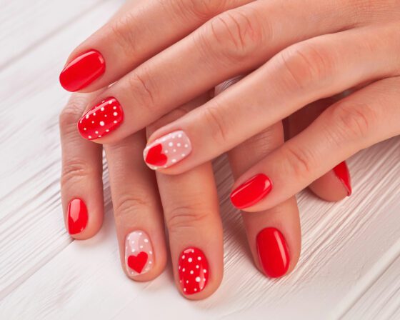 Close view of overlapping hands against pale wood, showing short bright red nails with white polka dots and pale pink accents with small red hearts