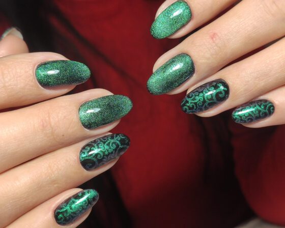 Close crop of both hands against a dark red backdrop, showing emerald green sparkle almond nails with dark accents carrying green scrollwork