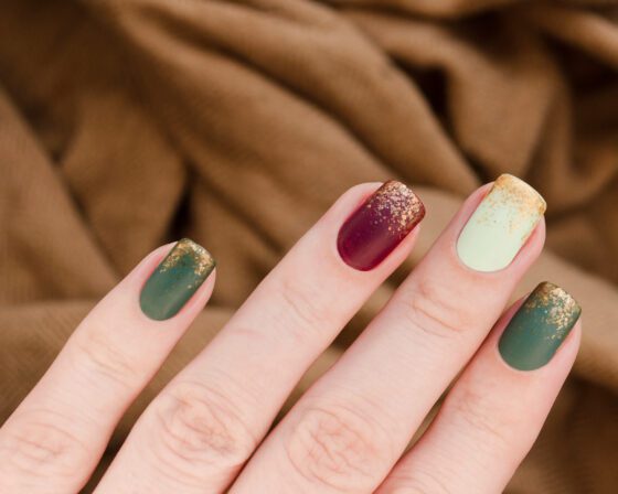 Close-up of a hand against warm brown knitted fabric, showing short matte nails in forest green, wine and cream with gold glitter at the tips