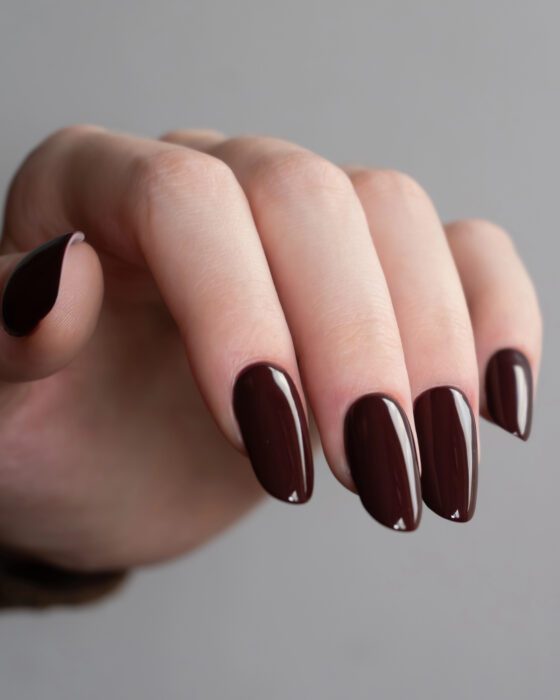Portrait close-up of downward-facing fingertips on a gray background, showing medium almond nails in a glossy deep burgundy shade
