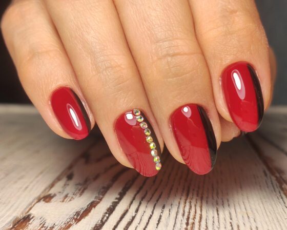 Close-up of one hand on a pale wood tabletop, showing glossy red nails with curved black side bands and a line of small crystals on an accent nail