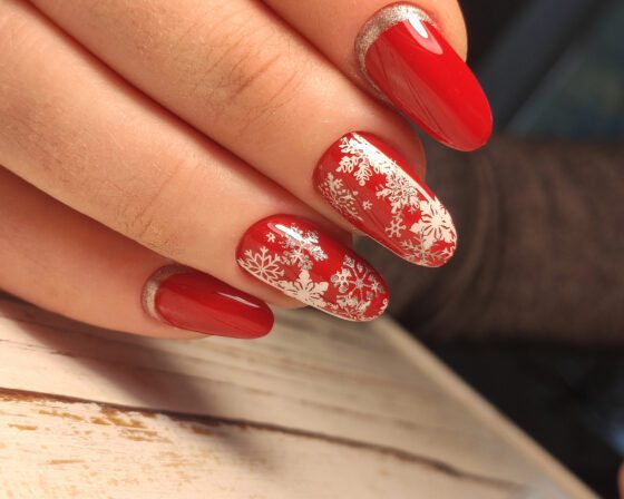 Macro view of fingertips on a pale wood surface, showing medium glossy red nails with white snowflake patterns on accents and silver sparkle near the base