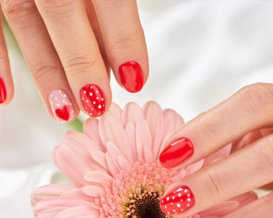 Tight close-up of fingers with a pale flower behind, showing short bright red nails with white polka dots and pale pink accents with small red hearts