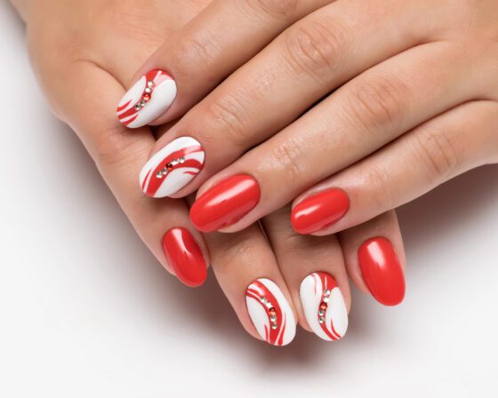 Tight close-up of crossed hands on a bright neutral background, showing glossy red nails and white accent nails with red swirls and small crystals