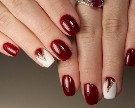 Close view of crossed hands against a neutral background, showing glossy burgundy nails with a white accent nail outlined by a fine gold V