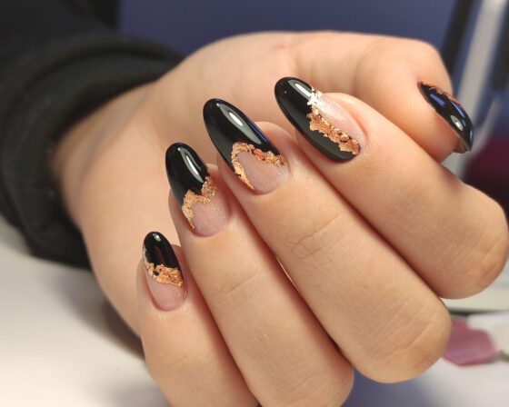 Tight close-up of a hand against a dark blurred background, showing glossy black and sheer nude almond nails edged with metallic gold flakes