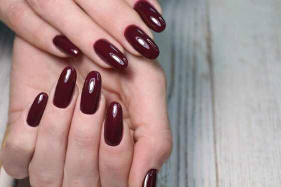 Close crop of overlapping hands against a softly blurred gray surface, showing medium-length nails in a single glossy deep burgundy shade