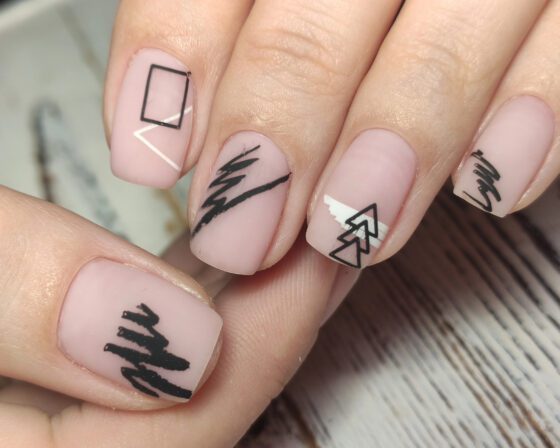 Macro close-up of fingertips over pale wood, showing short matte blush nails with black brush marks, outlined squares and fine white lines