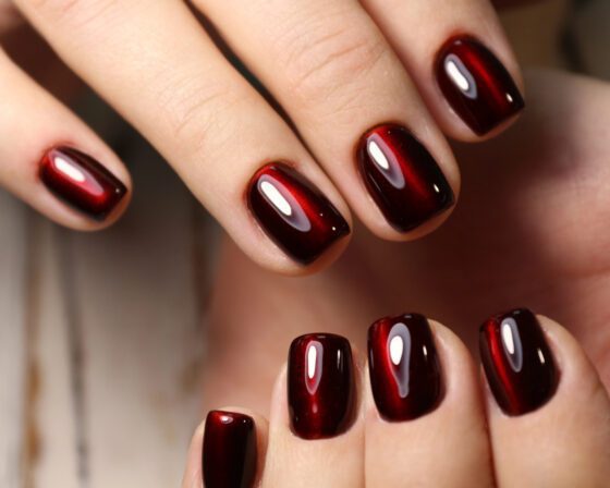 Macro view of fingertips against a softly blurred neutral surface, showing short squared nails in black cherry with a luminous red centre