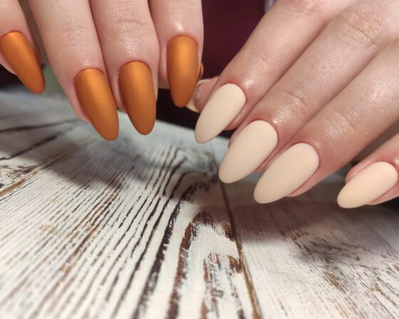 Two hands posed diagonally over pale wood and burgundy fabric, showing medium matte almond nails alternating metallic copper and cream