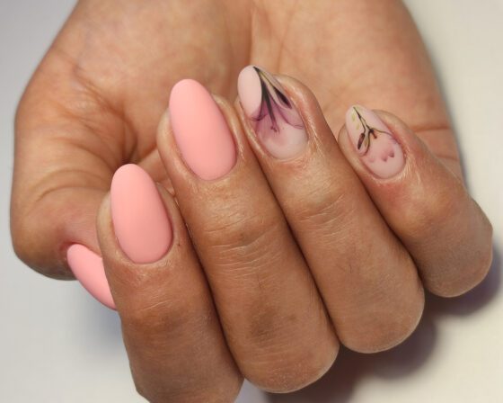Close view of curled fingers against a light neutral backdrop, showing matte peach almond nails with fine dark botanical sketches on sheer accent nails