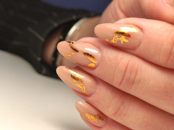 Macro crop of overlapping fingers with a dark prop at the edge, showing medium sheer nude almond nails with irregular metallic gold flakes