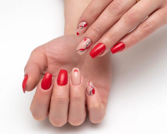 Two hands cupped together against a pale neutral background, showing bright red nails with nude accent nails carrying fine silver curves and tiny red hearts