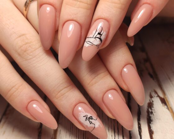 Hand fills frame over pale wood, showing long glossy peach-nude almond nails with small black line drawings on accent nails