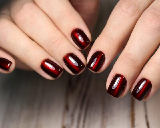 Tight close-up of multiple hands against a dark background, showing short squared nails in black cherry with a luminous red centre