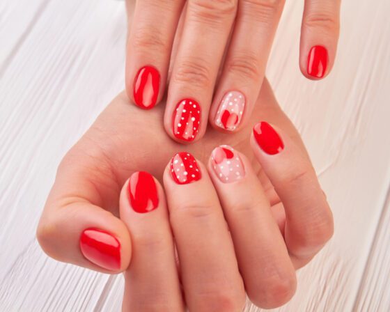 Close composition of overlapping hands against white, showing short bright red nails with white polka dots and pale pink accents with small red hearts