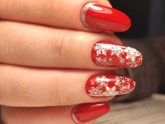 Macro close-up of fingers above a pale wood surface, showing medium glossy red nails with white snowflake patterns on accents and silver sparkle near the base