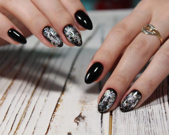 Two hands on pale wood with dark neutral background, showing glossy black nails with reflective silver flakes on accent nails