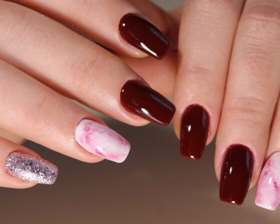 Close-up of crossed hands against a light neutral background, showing glossy burgundy nails with a pink-and-white marble accent and a silver glitter accent