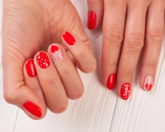 Multiple hands around a pale pink flower against a bright background, showing short bright red nails with white polka dots and pale pink accents with small red hearts