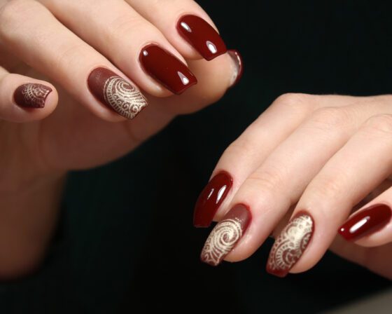Tight close-up of overlapping hands against dark fabric, showing glossy burgundy nails with cream and gold scrollwork on accent nails