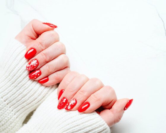 Two hands in a close studio pose with knit sleeve and open pale background, showing bright red nails with white snowflakes and fine sparkle on accent nails