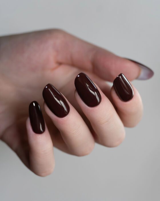 Portrait macro of curled fingers against a neutral gray background, showing medium almond nails in a glossy deep burgundy shade