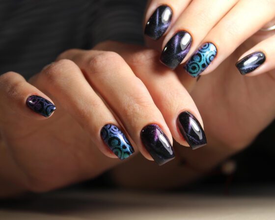 Layered close-up of several hands against dark blurred fabric, showing dark navy-violet shimmer nails with turquoise circle patterns on accent nails