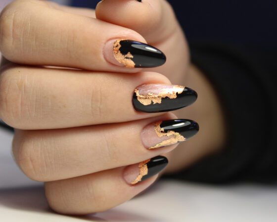 Tight horizontal crop of curled fingers against dark fabric, showing glossy black and sheer nude almond nails edged with metallic gold flakes