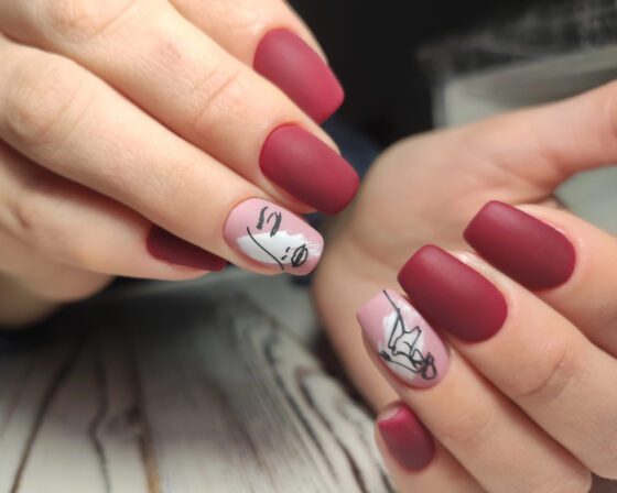 Close-up of overlapping hands above pale wood with a dark blurred background, showing matte berry-red nails with pale pink accents carrying black line drawings
