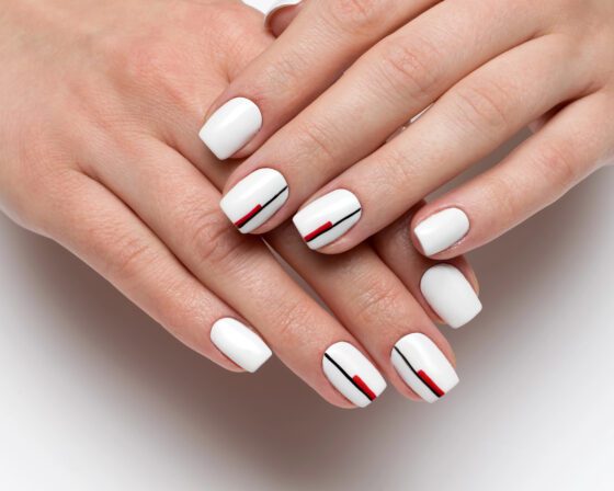 Close-up of crossed hands on a bright neutral background, showing short glossy white nails with thin red and black stripes on accent nails